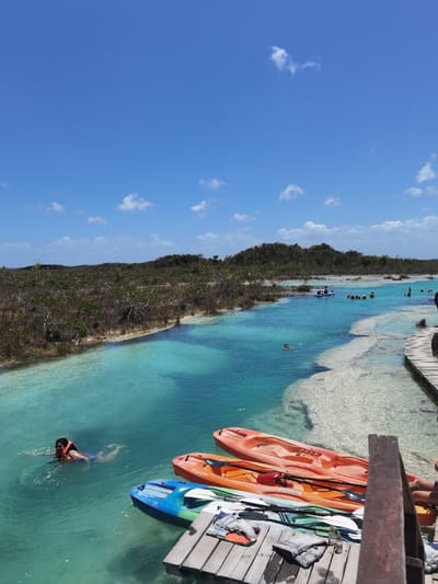 The image shows a view of a lagoon in Unidad de Riego, Mexico. It is a clear, bright, sunny day. The lagoon water is a brilliant turquoise blue. Three kayaks are docked on the white sand of the shore. A young man is swimming toward the kayaks. Another man is sitting on the wooden dock, which is at the edge of the water. The lagoon is bordered by trees and vegetation. There are a few other people in the lagoon, swimming and in kayaks in the distance. The scene is peaceful and tranquil. The sky is a bright blue with some wispy white clouds. The coordinates for the location are 18.591 latitude and -88.434 longitude.