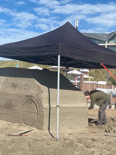 The image depicts an outdoor scene in Overveen, Netherlands, under a blue sky with scattered white clouds. A large, unfinished car-shaped sculpture, constructed from compacted earth or sand, occupies the foreground, featuring a circular relief detail on its side. A black pop-up canopy tent, supported by metal poles, shades the sculpture and work area; a red tie-down strap is visible on the right. Under the tent, one adult male, dressed in an olive-green jacket, dark grey cargo pants, and a dark beanie, is actively shaping the sculpture, bent over with his hands near its surface. Several hand tools, including a trowel, and red guide lines are visible on the sandy ground near the sculpture's base. In the midground, a vegetated sand dune rises, behind which multiple white patio umbrellas are visible. The distant background reveals a modern multi-story building with an angular roof and large windows, with a white telecommunication tower featuring antennas visible on or near its structure.