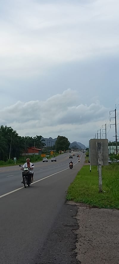 The scene depicts a road in Ban Thung, Thailand, viewed from a roadside angle. A person on a motorcycle is traveling on the right side of the road. Further down the road, several vehicles, including motorcycles and cars, are also in motion. On the right side of the road is grassy terrain and a concrete barrier. In the distance are trees and a mountain range under a cloudy sky. Power lines are visible overhead. Road signs are placed along the side of the road.