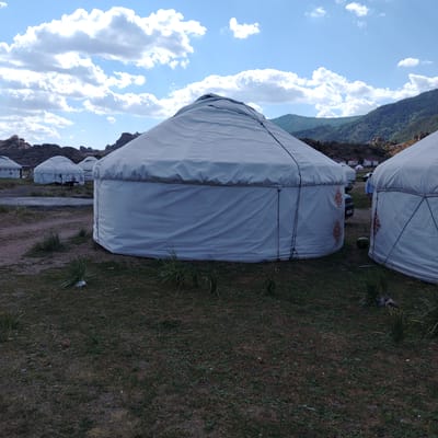 The image depicts a collection of yurts, traditional Mongolian dwellings, situated in an outdoor environment. The yurts are predominantly white with some displaying red decorative patterns on their exterior. They are arranged in a cluster, with some in the foreground and others further in the background. The ground around the yurts is a mix of dry grass and dirt. In the background, rolling hills covered with greenery and rocky outcrops are visible, under a partly cloudy sky with a clear blue sky dominating. A few vehicles, including a car, are parked near the yurts. There are also a few people visible, one of whom is standing between two yurts in the middle ground, facing away from the camera. The overall scene suggests a rural or nomadic settlement, possibly a tourist campsite, in the region of Торайғыров, Kazakhstan.