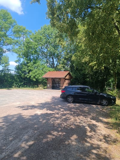 A black station wagon is parked on a gravel lot next to a small, open-sided brick shelter with a red tile roof. The shelter has a wooden support structure and appears to be a resting area. The lot is surrounded by lush green trees and foliage under a clear blue sky with scattered white clouds. The scene is illuminated by bright sunlight, casting dappled shadows from the trees onto the gravel. This location is near the Schloss Nordkirchen in Nordkirchen, Germany, a region known for its historic castles and gardens. There are no people visible in the image.