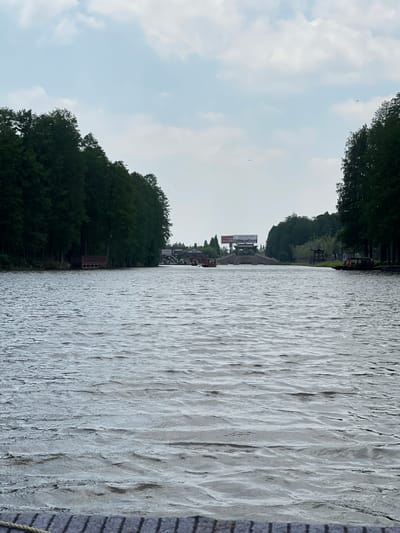 The image shows a body of water, likely a lake or wide river, with trees lining the opposite bank. The water surface has visible ripples. On the right side of the water, a billboard is visible in the distance. The sky is overcast with clouds. This image is taken in Xinghua, China.