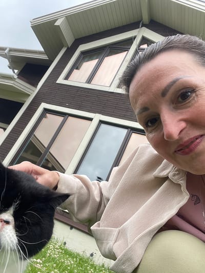 A medium shot captures a woman and a black and white cat outdoors, with a house in the background. The woman is in the foreground, facing the camera and holding the cat. Her face is visible, and she has dark hair pulled back, with makeup on. She is wearing a light-colored jacket over a pink shirt. The cat is black with white markings on its face and chest, and it is being petted by the woman. The house behind them has a dark brown, brick-like exterior and features large windows. The scene appears to be in a residential area, possibly in the city of Solov'evka, Russia, given the architecture and vegetation. The sky is overcast.