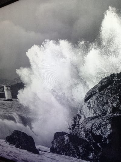 A black and white photograph captures a dramatic seascape. A large wave crashes against dark, jagged rocks in the foreground. Behind the wave, a small, white structure resembling a lighthouse stands on a low-lying island amidst turbulent waters. The sky is overcast with ominous clouds, enhancing the sense of raw power and untamed nature.