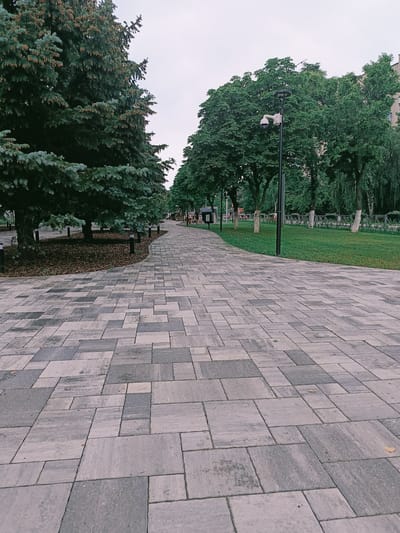 The image depicts a paved pathway in a park-like setting in Gubkin, Russia. The pathway is constructed from interlocking rectangular stone pavers in varying shades of grey. Tall evergreen trees line the left side of the pathway, with their branches extending over the paving stones. On the right side of the pathway, there is a well-maintained green lawn bordered by a decorative metal fence. Several trees with lush green foliage are planted along the lawn. A modern lamppost with a surveillance camera is visible in the middle ground on the right side of the pathway. The sky is overcast, suggesting a cloudy day. In the distance, along the pathway, a few indistinct figures can be seen walking, indicating public use of the area. The overall scene conveys a sense of urban green space designed for recreation and pedestrian use.