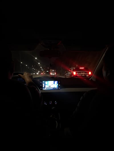 The image shows the interior of a vehicle at night, seemingly in slow-moving traffic in San Francisco Totimehuacán, Mexico. The vehicle is stationary or moving slowly, as evidenced by the illuminated brake lights of the car directly ahead, visible through the windshield. The driver is holding the steering wheel. A smartphone mounted on the dashboard is turned on, its screen displaying application icons. The dashboard controls are partially visible to the right of the phone. The overall ambient light is low, with the primary illumination coming from the external traffic lights and the smartphone screen.