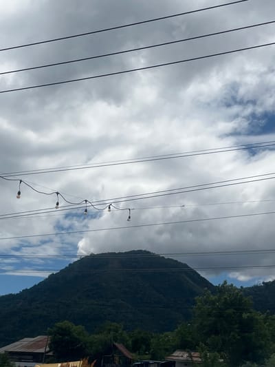 The image captures a scenic view of a mountain covered in lush green foliage under a cloudy sky. In the foreground, several power lines stretch across the frame, with a string of decorative lights hanging from one of them. Below the mountain, a few buildings with rusty roofs and dense trees are visible. The scene appears to be set in Panteraya, Indonesia, a region known for its natural beauty and mountainous terrain. The lighting suggests it is daytime, with diffused light filtering through the clouds. There are no people or animals visible in the image. The overall impression is one of a tranquil, rural landscape.
