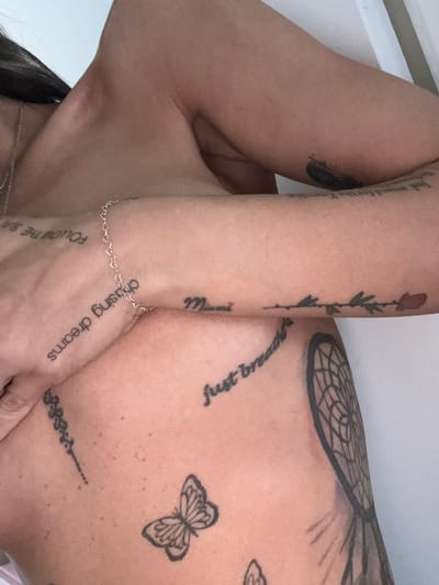 The image provides a close-up, angled view of a person's upper body, primarily focusing on their arms, shoulder, and chest, which are extensively covered in tattoos. The person's skin tone is warm, and the background is a plain, light color, suggesting an indoor setting.
On one arm, a tattoo of a red rose with a long stem and green leaves is visible, and below it, the word "Mama" is inked. On the chest and shoulder area, a large dreamcatcher tattoo with intricate webbing is prominent, alongside the phrase "Just breathe" written in a flowing script. Below these, a clear outline tattoo of a butterfly is visible. Another tattoo, appearing to be an abstract linear design, is situated further down on the body.
On the hand or lower forearm, the phrase "chasing dreams" is tattooed, and a partial view of "Follow the sun" can also be seen. A delicate silver bracelet made of small heart-shaped links adorns one wrist, and a thin silver necklace chain is partly visible around the neck.
The activity taking place is a pose to showcase the body art and jewelry. No specific time of day, weather, or emotional state can be determined from the image. While the location context of "Seal Beach, United States of America" is provided, there are no visual cues within the image itself to confirm this.