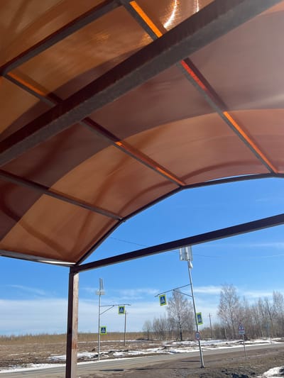 The image shows a view from under a metal and translucent orange polycarbonate structure, possibly a bus stop or shelter, in Gory, Russia. The photograph is taken at an angle, with the ground rotated 90 degrees counterclockwise. Outside the structure, the scene depicts a clear blue sky with some clouds, along with multiple pedestrian crossing signs and a speed limit sign (50). A secondary road intersects the main road with a corresponding road sign (100). There are power lines overhead. Beyond the road signs, there is sparse, dry vegetation and bare trees lining the roadside. Patches of dirt and possibly old snow can be seen near the road.