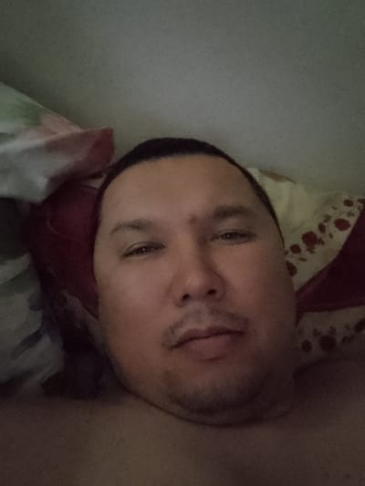 The image is a close-up, front-facing selfie of a person lying down, likely in bed. The subject is an adult male with medium skin tone, dark hair, and a round face. He has visible eyebrows, eyes, a nose, and lips. The background consists of pillows with a floral pattern. The lighting is dim, suggesting an indoor setting. The scene is consistent with a personal photograph taken in a domestic environment within Махамбет ауылдық округі, Kazakhstan.