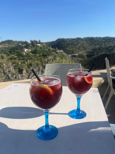 Two wine glasses, each filled with a dark red beverage, ice cubes, an orange slice, and a black straw, are centrally positioned on a light-colored, possibly wooden, outdoor table. Both glasses feature transparent bowls supported by bright blue stems and bases. Strong sunlight from the left casts distinct shadows of the glasses and other unseen objects onto the table surface. In the immediate background, parts of two light-colored outdoor chairs with decorative geometric patterns on their backs are visible. Beyond the terrace, a panoramic view unfolds under a clear, bright blue sky. This view encompasses undulating green hills, prominent rocky cliffs, and a winding river valley, consistent with the Tagus River's course. Several distant buildings, characterized by light-colored walls and reddish roofs, are sparsely visible within the natural landscape. The scene is situated on an outdoor terrace of an establishment in Toledo, Spain, overlooking the Tagus River valley and its surrounding natural environment.