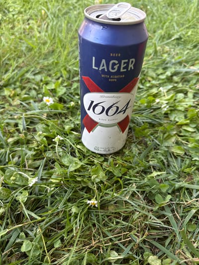 This is a close-up, horizontal shot of an opened can of Kronenbourg 1664 Lager beer lying in the grass. The can is mostly blue with white and red accents. The text "LAGER" is prominently displayed in large white letters, with "BEER" above it and "WITH ALSATIAN HOPS" below. The Kronenbourg logo and the number "1664" are also visible. The can appears to be condensation-covered, suggesting it is a cold beverage.

The can is positioned diagonally on a bed of green grass and small white wildflowers. The grass is lush and appears to be slightly damp. The overall scene evokes a relaxed, outdoor setting, possibly a picnic or a casual gathering in a park or garden. The lighting suggests it is daytime, with soft natural light illuminating the scene. There are no people visible in the frame, focusing entirely on the discarded beverage and its natural surroundings. The presence of the opened can hints at an activity that has recently concluded. The location context of Longpont-sur-Orge, France, adds a specific geographic detail to the scene, but the visual cues within the image itself are more general.