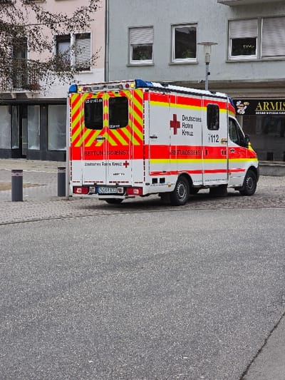 A white German Red Cross (Deutsches Rotes Kreuz) ambulance is parked on a cobblestone surface in Zweibrücken, Germany, specifically in front of Armisan Kebab & Pizza Haus at Hauptstraße 140. The ambulance displays a white body with prominent red and yellow reflective striping, the red cross emblem, and "RETTUNGSDIENST" (Rescue Service) text on its sides and rear. The emergency number "112" is visible on the passenger side, and the license plate "ZW GR 831" is affixed to the rear. Blue emergency lights are mounted on the vehicle's roof.

The scene features a dark gray asphalt foreground leading to the cobblestone area where the ambulance is situated. In the background, a multi-story gray building with several rectangular windows, each fitted with white venetian blinds, houses the "ARMISAN KEBAB & PIZZA HAUS" on its ground floor, indicated by a visible sign. To the left of this building, another structure with a light-colored upper facade and dark ground-floor windows is partially visible, with a bare tree branch extending into the frame from its vicinity. Two dark bollards are positioned on the cobblestone near the ambulance's rear, and a street lamp stands between the ambulance and the gray building. No individuals or ongoing activities are visible.