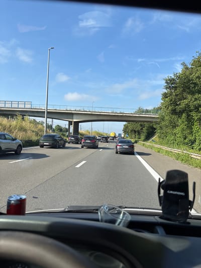 The image is taken from the perspective of a vehicle driver on a highway, likely in or near Vilvoorde, Belgium, given the presence of a street sign with what appears to be Belgian license plates and European highway infrastructure. The scene depicts moderate traffic with several cars, mostly black SUVs and sedans, driving on multiple lanes. An overpass crosses the highway in the mid-ground, with grassy verges and trees lining the sides. The sky is clear blue with some wispy clouds. In the foreground, on the dashboard of the vehicle, is a red can and a phone holder with the brand "GRUNDIG" visible. The overall impression is that of a normal daytime commute on a highway.