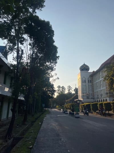 The image shows a paved road in Pekanbaru, Indonesia, with a building that appears to be a mosque on the right side of the frame. The building is light-colored with arched windows and a decorative dome. A line of manicured bushes runs along the side of the building. On the left side of the road, there is a building with a tiled roof and a tree-lined area with a drainage ditch. Two people are seen riding motorcycles on the road. The sky is clear and bright, indicating it is daytime. The overall scene suggests a well-maintained urban or campus environment.