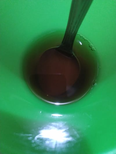 A spoon is partially submerged in a brown liquid within a green bowl. The spoon's handle is visible at the top of the frame, and its bowl is immersed in the liquid, obscuring the bottom of the bowl. The liquid appears to be tea or coffee, with some small bubbles forming on its surface near the spoon. The green bowl has a smooth, slightly reflective interior. There are some bright reflections on the surface of the liquid and the bowl, suggesting overhead lighting. The overall scene is a close-up shot, focusing on the contents of the bowl. There is no indication of the location being Gampongbaro, Indonesia.