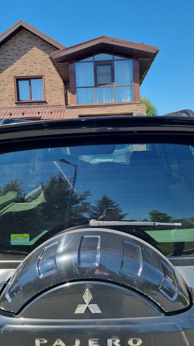 In the city of Verzilovo, Russia, a black Mitsubishi Pajero is parked in front of a two-story brick house with a brown roof. The house has multiple windows, including a large bay window on the second floor. The car's rear window is visible, reflecting the sky and trees. A license plate with the characters "C07900 799" is affixed to the car. The spare tire cover, emblazoned with the Mitsubishi logo, is prominent on the back of the vehicle. A caution sticker is visible on the lower left side of the rear window. The sky is clear and blue.