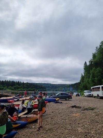 The image depicts a rocky riverbank near the city of Visherogorsk, Russia, on an overcast day. A group of people are gathered around several inflatable rafts, some of which are loaded onto the shore. A dark sedan with its hood open is parked near a tent, and further down the bank, a white van and a white bus are visible. The river is calm and bordered by a dense forest of green trees on the opposite bank. Several individuals are seen standing, sitting, or interacting with the rafts. One person is wearing a camouflage jacket and shorts, while another is wearing a yellow jacket and black pants. The overall scene suggests a recreational outing or a staging area for water activities.
