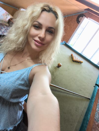 The image depicts a woman inside a casual indoor setting in Терновский округ, Ukraine. A blonde woman with curly hair and blue eyes is captured in a selfie, smiling towards the camera. She wears a light blue, white-patterned, spaghetti-strap dress. Two gold-colored chain necklaces, one with a circular pendant, are visible around her neck. Behind her, to the left, is a floral-patterned pillow and what appears to be part of a bed or seating area. The right side of the frame features a green-felted billiard table with a rack of fifteen brown wooden balls arranged triangularly at one end, and a single brown wooden ball positioned separately closer to the viewer. A wooden cue stick lies across the table. In the upper right background, a bright window indicates daylight, alongside wooden wall paneling and a decorative circular object mounted on the wall. Wooden beams are visible near the ceiling.