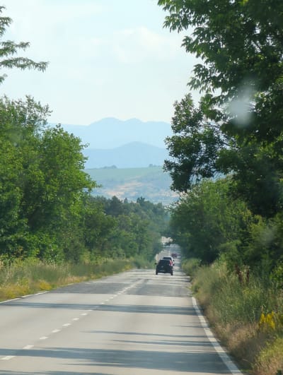 The image shows a road with several cars driving on it, bordered by lush green trees and vegetation. The road appears to be a two-lane highway with a white line marking the center and dashed white lines indicating the lanes. The trees create a canopy over the road in some sections, casting shadows on the asphalt. In the distance, rolling hills and mountains are visible under a bright, clear sky, suggesting a sunny day. The scene evokes a sense of travel and a journey through a scenic, possibly rural, landscape. The orientation of the image is vertical, giving a sense of looking down from a slightly elevated perspective or a tilted camera. No people are clearly visible, but the presence of cars indicates people are traveling. The overall mood is serene and peaceful, characteristic of a road trip through nature. The location context suggests Ohoden, Bulgaria, which aligns with the mountainous terrain and vegetation depicted.