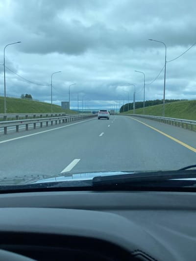 The image shows a view from inside a vehicle looking out onto a highway. The sky is overcast with heavy clouds. The highway has multiple lanes, separated by metal guardrails and white and yellow lane markings. Streetlights line the side of the road. On the right side of the highway, there is a grassy embankment. In the distance, there are a few vehicles, including a white SUV and a truck. The foreground of the image is dominated by the dashboard and windshield wipers of the vehicle. The image appears to be taken in a rural or suburban area, likely on a highway.