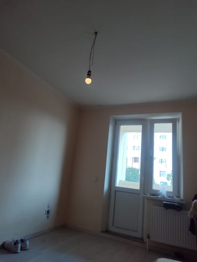The media file shows an interior scene, likely a room undergoing renovation or recently completed. The walls are painted a light, neutral color, and the ceiling is white with a subtle crown molding. In the center of the ceiling, a single lightbulb hangs from wires, illuminated, suggesting it's either the only light source or being used for temporary lighting.

To the right of the frame, there is a large double window or door with white frames, offering a view of buildings and trees outside, indicating an urban or suburban environment. Below the window, a white radiator is visible. On a windowsill above the radiator, a few objects are placed, including what appears to be a black box and some white clutter, possibly cleaning supplies or renovation materials.

On the left side of the room, a pair of white sneakers is placed on the floor near the corner. The floor appears to be light-colored laminate or wood. A white electrical outlet is visible on the wall near the sneakers. The overall lighting suggests it is daytime, though the exact time is not discernible. The scene is devoid of people, giving a sense of emptiness or quiet. The setting appears to be a residential room in Rezina, Moldova, judging by the provided location context. No visible text is present in the image.