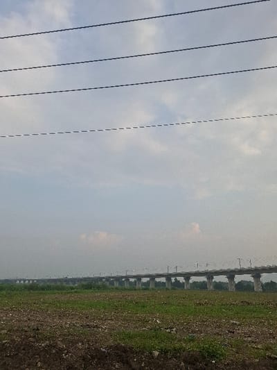 The image shows a concrete overpass with a light haze in the background. There is a grassy field in the foreground and a cloudy sky overhead. The overpass is located in Kecamatan Cimahi Selatan, Indonesia, at coordinates -6.923 latitude and 107.546 longitude. No people or objects are visible in the image. There are power lines running across the top of the image. The overpass is made of concrete pillars and beams, and there are several supports running underneath it. The grass in the foreground is green and sparse, with a few patches of dirt. The sky is a light blue color, with some clouds. The overpass is constructed of concrete. The ground is mostly dirt and has sparse, short grass growing on it. The photo was likely taken from a distance, as the overpass appears quite small in the image. The subject of the photo is the overpass. The overpass is the only object in the image that is in focus. The image is likely of the elevated portion of the Bandung Raya commuter line, which is a light rail line that connects Bandung and its suburbs. The line opened in 2015. The image is in focus and well-lit. The photo was taken in the late afternoon or early evening, as the sun is low in the sky.