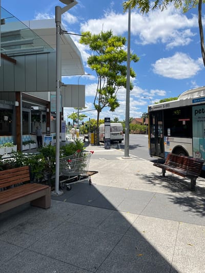 This media file captures a bright, sunny daytime street scene in Indooroopilly, Australia, featuring an urban commercial area. The image is oriented vertically (rotated 90 degrees clockwise), showing a sidewalk alongside a road with buildings and greenery.

In the foreground, a wide, tiled sidewalk is visible, with distinct shadows cast across it by the bright sun. A modern wooden bench is situated on the right side of the sidewalk. Further along the path, a silver shopping cart with red handles is positioned near some green foliage.

To the left, contemporary buildings with reflective glass panels and light-colored facades are visible, housing businesses. A person can be seen standing near a shop entrance. Lush green trees line the street, providing natural elements to the urban landscape.

A prominent feature is a white and blue public transport bus, stopped or parked on the road. Visible text on the bus includes its identification "S 1951", the instruction "Get on board", "EMERGENCY EXIT", and a digital display showing "437". Fragments of advertisements such as "petrol", "We hustle so you can...", and "PRICE BEAT" are also discernible on its side. A metal bus stop bench is located near the bus. Further down the street, a white van, traffic lights, and other street infrastructure are present. The sky above is a vibrant blue with scattered white, puffy clouds. The scene suggests everyday activity in a lively commercial district under clear weather conditions.