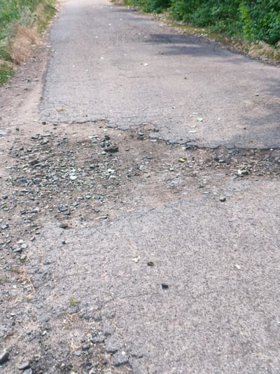 The image shows a section of a rural asphalt road with visible damage and debris. The road surface is cracked and appears worn, with several potholes. The shoulder of the road on the left side is unpaved and covered in gravel and dry vegetation. On the right side, dense green foliage borders the road. Scattered across the asphalt are small pieces of light-colored debris, possibly plastic or glass fragments, and a few scattered leaves. The scene depicts a neglected or weathered roadway in a natural setting, likely outside of a densely populated urban area within the city of Psilske, Ukraine. No people or vehicles are visible in the frame.