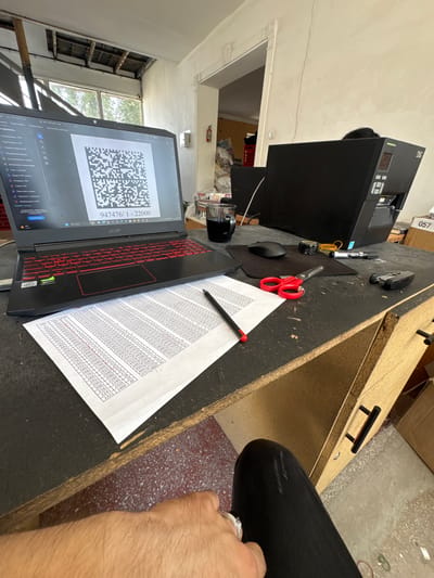 A medium shot captures a cluttered, work-in-progress desk environment, suggesting an office or workshop setting, possibly in Danghara, Uzbekistan, given the context. A black Acer laptop, with its distinct red-backlit keyboard, is open, displaying a QR code and the text "947476/1 - 22000" below it. The laptop screen is angled towards the viewer, and the laptop itself sits atop a black mousepad. To the right of the laptop, a black desktop computer tower is prominently placed, with a visible "TSC" logo on its upper right corner. A glass mug containing a dark liquid, likely coffee or tea, rests to the left of the laptop.

In front of the laptop, a white sheet of paper with numerous columns of numbers and alphanumeric codes is spread out. A black pen with a red tip lies on this paper, positioned diagonally across it. A pair of red-handled scissors is placed on the paper to the right of the pen. Other desk items include a small roll of black tape, a marker pen, a stapler, and a black computer mouse.

The desk surface itself appears to be made of dark, worn material, with visible scratches and imperfections. The background reveals a slightly unfinished interior with exposed ceiling elements and a doorway leading to another room. A fire extinguisher is mounted on the wall to the left. The overall scene is one of active work or study, with materials spread out and tools readily accessible. The lower right of the image shows a person's left leg clad in black, ripped jeans, and a hand wearing a ring, suggesting the photographer's presence at the desk. The floor is a concrete surface with scattered debris.