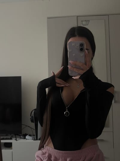 An indoor scene in what appears to be a residential room in the city of Krasnaya Zarya, Russia, is depicted. A single individual, a female, is visible from the chest down, reflecting in a mirror while holding a smartphone. She has long, dark hair parted in the middle. She is wearing a long-sleeved black bodysuit with an open zipper extending to below the chest, revealing a silver pendant worn around her neck. The bodysuit features thumbholes on the sleeves and cut-outs on the shoulders. Below the bodysuit, she wears light pink elastic-waist bottoms, possibly sweatpants. Her left hand is raised, with fingers splayed, near her collarbone. Her right hand holds a light purple smartphone with a bow-patterned case, obscuring her face and upper right chest. Her nails are manicured with light pink polish.
The background, visible around the individual's reflection, includes a light-colored wall on the left. On the far left, a dark-colored flat-screen television is positioned on a white console or cabinet. To the right of the television, a white gaming console (possibly a PlayStation 5) stands upright, with dark headphones resting near its base. Further to the right, a large, light-colored wardrobe or cabinet with reflective surfaces is visible, suggesting the mirror is integrated or placed against it.