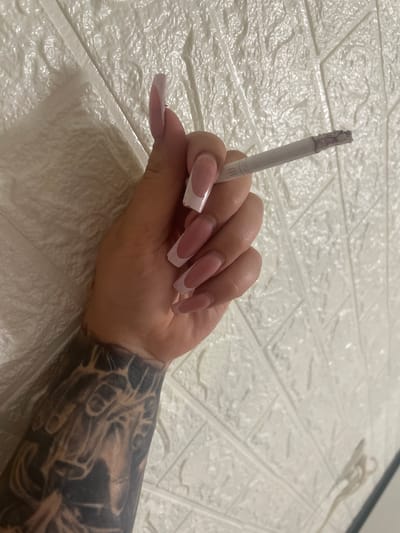 A close-up shot shows a person's right arm and hand holding a lit cigarette. The hand has long, manicured fingernails with a French tip design. The person's forearm is covered in a black and grey tattoo featuring various designs. The background consists of a textured white wall, possibly wallpaper. The image does not provide any information about the location in the city of Коробицыно, Russia.