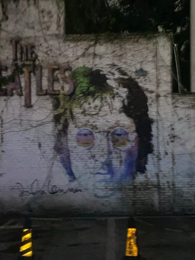 The image displays a piece of street art or a mural painted on a weathered, light-colored building wall that is heavily overgrown with dark green vines or ivy. The central subject of the artwork is a stylized portrait of John Lennon, recognizable by his iconic round spectacles and dark, somewhat shaggy hair. The art style appears expressive, featuring blue and purple paint drips extending downwards from the right side of his face. A patch of green color is also visible on the left side of his head, blending with the surrounding foliage.
To the left of the portrait, large, block-style letters are painted vertically on the wall, spelling out "BEATLES." These letters appear distressed, matching the aged texture of the wall. To the right of Lennon's face, a faint, handwritten signature, which seems to be "John Lennon," is visible.
The overall lighting is dim, suggesting the photograph was taken at night or in very low light conditions. On the far right side of the frame, bright, rectangular, yellow-orange light sources are visible, possibly emanating from windows or other architectural features. The wall itself shows signs of age and discoloration, contributing to the rustic, urban aesthetic of the scene.