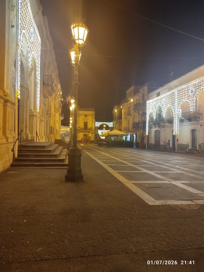 The image depicts a nighttime scene in a city square in Militello in Val di Catania, Italy. On the left, a building facade is decorated with intricate, illuminated patterns, possibly a historic building or a church. Adjacent to this building is a set of wide, stone stairs with handrails. A tall, dark lamppost with multiple lights stands in the foreground, illuminating the paved square. The square itself has a patterned design of light-colored squares on a darker paved surface. In the background, other buildings line the square, some also adorned with decorative lights. There are outdoor seating areas with umbrellas, suggesting a cafe or restaurant. One person, wearing a red shirt and white shorts, is visible walking away from the viewer on the right side of the square. Another person is partially visible near the outdoor seating area. The sky is dark, indicating nighttime. A timestamp "01/07/2026 21:41" is present in the lower right corner of the image.
