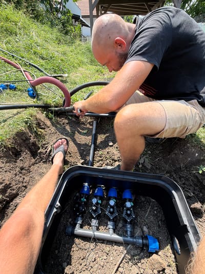 The image shows a man working on an irrigation system in a grassy outdoor area, likely a garden or yard, in Bašť, Czechia. The man, with a shaved head and beard, is wearing a black t-shirt and tan shorts. He is kneeling and appears to be connecting hoses or pipes to a black plastic irrigation valve box partially buried in the dirt. His left foot, wearing a flip-flop, is visible in the foreground, as is his left leg. Several pipes and hoses, including a red corrugated one and black flexible tubing, are present in the scene, along with blue connectors. The background consists of dense green grass and some vegetation. A portion of a building with a dark roof structure is visible in the upper right corner. The lighting suggests it is daytime with bright sunlight.