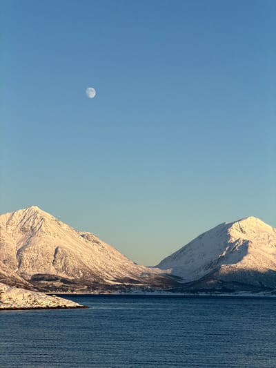 The image displays a clear daytime winter landscape located in Lødingen, Norway. The scene is composed of a light blue, cloudless sky filling the upper half of the frame, within which a crescent moon is distinctly visible in the upper left quadrant. The middle ground is dominated by two large, snow-covered mountains flanking a visible passage or valley. The left mountain presents significant snow coverage on its peaks and slopes, with areas illuminated by a low sun exhibiting a warm, golden coloration, contrasting with shaded sections. The right mountain is also extensively snow-covered, displaying similar lighting conditions with direct sunlight on its upper contours and shadows on its lower portions. The foreground features a calm body of dark blue water, consistent with a fjord, exhibiting minor surface ripples. A small, snow-dusted rocky landmass extends into the water from the immediate left foreground. No human figures, animals, or active vehicles are observed. The environmental context indicates clear weather conditions during a period of snow coverage, with a low sun angle suggesting either early morning or late afternoon.