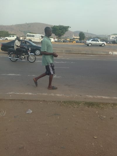 A man is walking along a paved road with a motorcycle driving past him. The man is wearing a light green shirt and black shorts. The motorcycle is black with a silver seat and white handlebars. The road is grey and has white lines down the center. There is a white car in the background of the image. The man is walking away from the camera. The motorcycle is moving towards the camera. The scene is located in Maraba, Nigeria, at the coordinates latitude: 9.024, longitude: 7.573. The road is relatively empty and is bordered by a dirt shoulder. The image is taken from a low angle.
