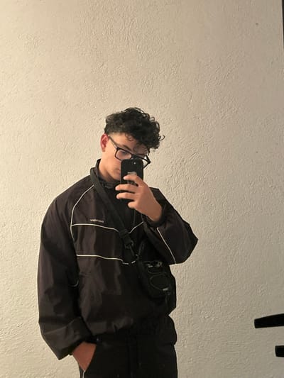 This image depicts a male individual taking a mirror selfie inside a location in Hvostiane, Bulgaria. The individual has dark, curly hair and wears black-rimmed eyeglasses. He is dressed in a black track jacket with two horizontal white stripes across the chest and white piping along the sleeves. A reflective logo, appearing as "adidos" due to the mirror reflection, is visible on the left chest area. He is also wearing a black crossbody bag with a smaller black pouch attached to the strap. The individual holds a dark-colored smartphone with his left hand, positioned to capture his reflection. His right hand is partially obscured, appearing to be in a pocket. The background consists of a plain, light-colored, textured wall. A dark, indistinct object is visible in the lower right corner of the frame.