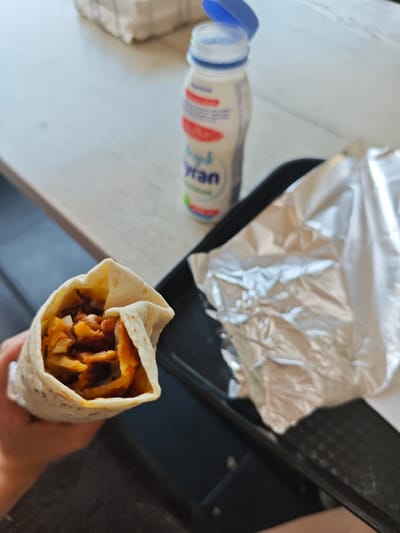 A hand holds a partially unwrapped shawarma, revealing seasoned meat, fries, and white onions inside a flatbread wrap. A plastic bottle of Ayran yogurt drink with a blue lid sits to the right, next to a piece of aluminum foil on a black tray. The scene is set on a light-colored wooden table. The provided crops do not contain enough visual information to determine the specific location within Tilburg, Netherlands.