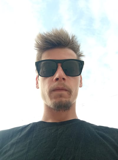 A low-angle, waist-up, outdoor, selfie-style photograph shows a light-skinned male wearing dark sunglasses and a black t-shirt, with his blond hair spiked upwards. He has a mustache and a goatee. The background consists of a bright sky with sparse clouds, suggesting it is daytime. The image appears to have been taken in Birži, Latvia.
