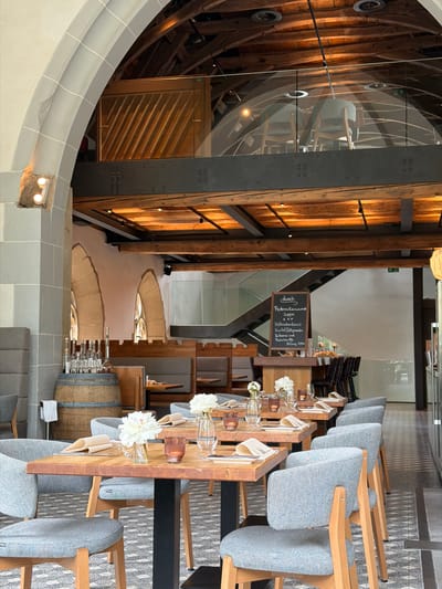 This image captures a restaurant interior in Bad Sobernheim, Germany. The building has a unique architectural style with exposed wooden beams and steel supports. There is a menu on a small blackboard displaying "Dunch" and a list of dishes. Tables are set with flowers, glassware, and napkins. Wooden chairs with gray fabric upholstery are arranged along the tables. A large wooden barrel is partially visible near a set of tables. Arched windows are seen in the background, suggesting a historical building. The overall lighting is warm, creating a pleasant ambiance.