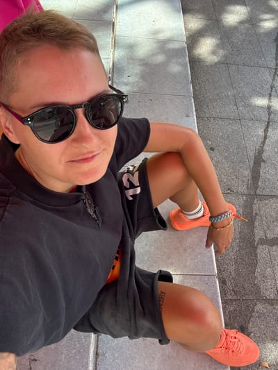 This is a high-angle, medium shot of a person lying down outdoors. The person is wearing a black t-shirt, grey shorts, and bright orange sneakers. They have short blonde hair and are wearing black sunglasses. One arm is bent, resting on their leg, with a bracelet on their wrist and a gold chain necklace visible. Their legs are crossed at the knees. The ground beneath them appears to be a sidewalk with visible tiles and some shadows.
The setting suggests a casual, daytime activity, possibly relaxing or resting outdoors. The lighting indicates it's daytime, with shadows suggesting sunlight. The person's relaxed pose and casual attire contribute to a laid-back mood.
There is some visible text on the person's clothing, a white "12" on the sleeve of the t-shirt, and a tattoo on their leg that reads "RECOVERY" in black ink, though it's partially obscured.
The image does not contain any explicit cues to indicate the specific time of day or weather conditions beyond the presence of sunlight. The overall impression is one of casual relaxation in an urban outdoor environment.