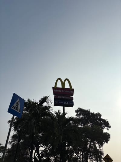 The image shows a low-angle view of a McDonald's sign and a pedestrian crossing sign against a hazy sky. The McDonald's sign is yellow and iconic, with "McDonald's" written below it. Underneath the McDonald's sign are two smaller signs: one reads "DRIVE THRU" and the other, in Indonesian, indicates "Buka 24 jam," meaning "Open 24 hours." A blue pedestrian crossing sign, showing a white silhouette of a person walking on a crosswalk, is positioned to the left of the McDonald's sign. Silhouetted palm trees and other foliage are visible in the foreground, partially obscuring the lower parts of the signs and pole. The sky is a pale blue-grey, suggesting either early morning or late afternoon light, or possibly atmospheric haze. The overall scene is urban, likely a roadside location in Pakulonan Barat, Indonesia, given the presence of Indonesian text on one of the signs. No people are clearly visible, and no specific actions are depicted. The environment appears to be outdoors, near a road.