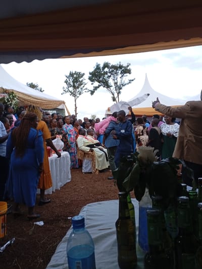 An outdoor gathering, possibly a celebratory event, is captured from under a tent, providing shade for attendees. The scene is set on reddish-brown earth, likely dirt or gravel, consistent with a rural or semi-rural setting in Shimbi Mashariki, Tanzania. The sky above is overcast, suggesting a cloudy day.
A large crowd of people, dressed in a variety of colorful attire, including patterned fabrics, are gathered. Many are standing and observing the central area. In the mid-ground, an older man, appearing to be the guest of honor, is prominently seated on an ornate golden chair, wearing a light-colored robe with gold trim. To his left, a white table draped with a white tablecloth is visible, and people are standing nearby.
In the immediate foreground, a table is laden with bottles, including several green and brown glass bottles (likely beer bottles), a plastic water bottle containing a blue liquid, and a single white rose with green leaves placed in a vase or bottle. A yellow bucket rests on the ground to the left.
The atmosphere appears festive and positive, with a man on the far right giving a thumbs-up gesture. Several white party tents, some decorated with gold fabric, are visible in the background, along with a few sparse trees. No clear text is discernible in the image. The event seems to be a significant social occasion, possibly a wedding or a traditional ceremony, given the number of attendees and the setup.