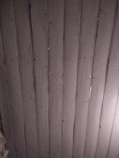 The image shows a close-up, low-angle view of a wooden ceiling with vertical planks. The wood is painted a dull, medium-brown color, and the planks have visible seams between them. The lighting is dim, with a light source appearing to be from the right side, casting subtle shadows and highlights along the wood grain and seams. The surface appears somewhat textured and possibly aged. There is a small amount of what looks like scattered debris or imperfections on the surface, along with a few small dark marks that could be knots or blemishes in the wood. The top left corner shows a hint of a patterned object, possibly upholstery or fabric, in muted colors. The overall impression is of an interior ceiling made of wooden planks. There is no visible indication of the city of Bandza, Georgia, in this image.