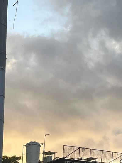 A low-angle, outdoor, medium shot captures a portion of a metallic utility pole in the foreground on the left, extending vertically from the bottom edge upwards. A dark, thin wire hangs from the top of the pole, extending diagonally into the sky. The sky dominates the upper two-thirds of the image, filled with a mix of light and dark gray clouds, with patches of pale blue sky visible at the top. In the lower portion of the image, the rooftops of buildings are visible. A large, white, cylindrical water tank is prominent on the left side of the rooftop. To the right of the water tank, a metal fence structure with a diamond pattern encloses an area on a rooftop. A small, open-sided shelter with a roof is also visible on the rooftop. The lower edges of green foliage are present at the bottom left of the frame. The scene appears to be set in Conuco Viejo, Venezuela, during daylight.