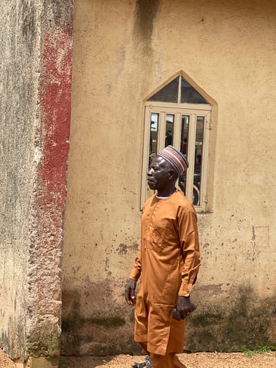 An older Black man wearing a brown traditional tunic and a striped cap stands in front of a weathered, tan-colored building with a unique triangular window. The building's wall has some dark stains and patches. To the left of the building is a rough, textured wall with a vertical red stripe. The ground is covered with small stones and gravel, with some sparse green vegetation. The man is looking to his left and holding a dark object in his right hand. Reflections in the window suggest other people and structures are present. The overall scene depicts an outdoor environment in Kassa, Nigeria.