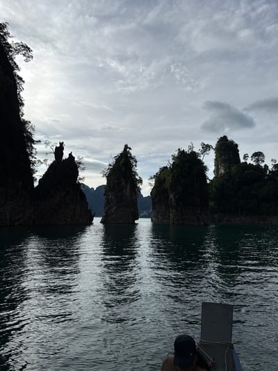 The image depicts a body of water with several karst formations rising from it. The formations are silhouetted against a cloudy sky and are covered in dense vegetation. In the foreground, the front of a boat is visible, with a person wearing a black cap looking down. The water is calm, with ripples reflecting the surrounding landscape. The scene is indicative of the dramatic limestone cliffs and lush greenery characteristic of Khao Phang, Thailand, a region known for its stunning natural beauty, particularly within Cheow Lan Lake. The lighting suggests it is daytime, with an overcast sky.