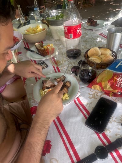 A table is set with a white tablecloth adorned with red stripes and floral patterns. A variety of food items including meat, salad, bread, and chips are scattered across the surface, alongside drinks and dining utensils. A person's hand wielding a fork is visible, suggesting someone is in the midst of eating. A mobile phone and a smart watch also sit on the table. The overall scene appears to capture a casual meal or picnic setting.