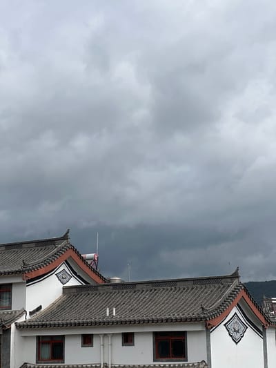 The image captures a view of traditional Chinese architecture in Eshan Yi Autonomous County, China, under a heavy, overcast sky. The foreground is dominated by the tiled roofs and white walls of several buildings. The roofs feature the characteristic curved eaves and grey tiles. Decorative elements are visible on the white walls, including a black and white pattern within a diamond-shaped frame on the upper gable. Red-framed windows are scattered across the facades. A solar water heater is visible on one of the rooftops. The sky is filled with dark, ominous clouds, suggesting an impending storm. In the background, a blurred hillside is visible. There are no people or discernible activity in the frame.